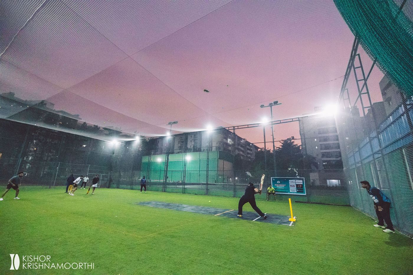 A floodlit box cricket ground at dusk